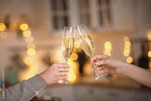 Two women clink glasses with champagne close-up of hands against the backdrop of a Christmas tree and Christmas lights. Lesbian couple celebrating. Women's hands hold thin crystal glasses against the 
