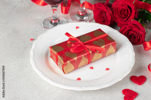 Gift in wrapping paper on white plate  red roses and two glasses for wine  concept for Valentines Day