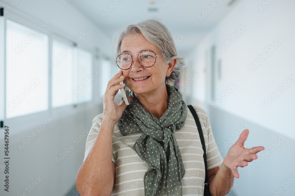Portrait of senior Caucasian woman walking in a corridor of a building while using the phone. Attractive elderly lady talking on smartphone gesturing Portrait of senior Caucasian woman walking in a corridor of a building while using the phone. Attractive elderly lady talking on smartphone gesturing