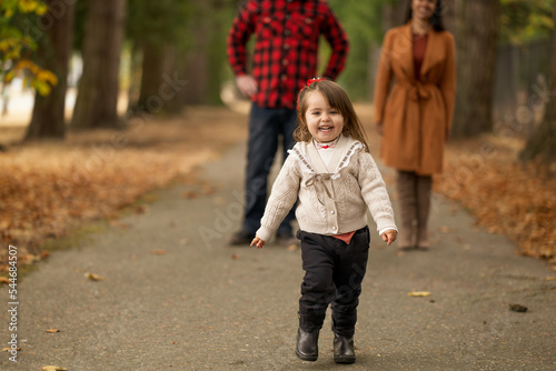 A cute little girl is playfully running away from her parents in a beautiful autumn colored park