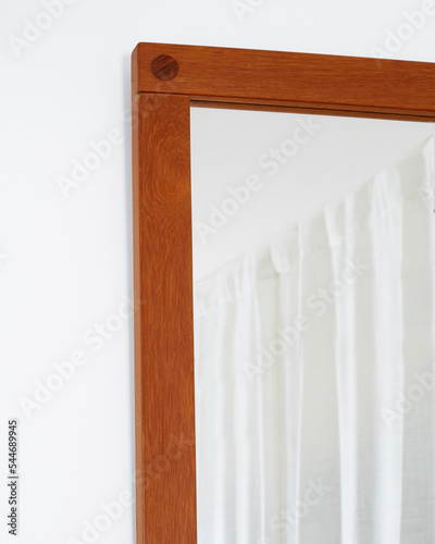 Vintage teak mid-century modern frame mirror. Warm brown wood mirror. Corner detail view with white wall and curtains. 