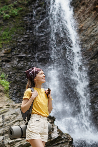 A young attractive female tourist in a yellow t-shirt and scarf is smiling and looking at the high waterfall behind her. Concept of bloggers  young activists. Place for your design