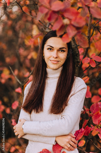 Portrait, photograph of a beautiful, young long-haired girl, brunette woman in an expensive beige suit in autumn against the background of red bushes, trees with leaves.