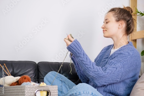 A young woman knits a hat. The woman is dressed in a sweater in the color of peri peri. The process of knitting a hat on knitting needles. Close angle