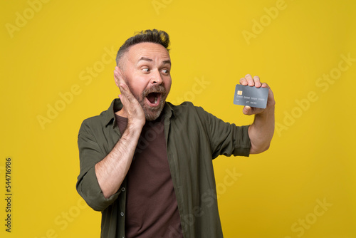 Excited mature man in casual green shirt posing isolated on yellow orange wall background looking on hand holding bank credit card, studio portrait. People sincere emotions lifestyle finace concept. photo