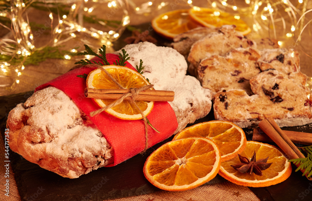 Fototapeta premium Christmas Stollen. Traditional Sweet Fruit Loaf with Icing Sugar. Xmas holiday table setting, decorated with garlands, baubles, walnuts, hazelnuts, cinnamon sticks.