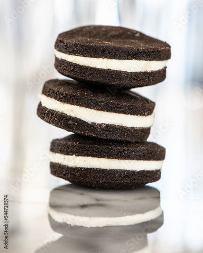 Close up of three chocolate sandwich cookies filled with white chocolate ganache frosting and stacked on top of each other, slightly askew.  Reflection of cookies shows in white marble slab. 