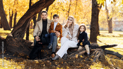 Happy family in an autumn park. Mother, father, son, daughter and dog are sitting on a tree trunk and looking into the camera, yellowed trees around
