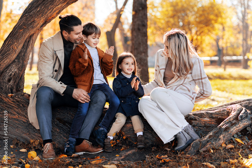 Happy family in an autumn park. Mother, father, son and daughter are sitting on a tree trunk, yellowed trees around