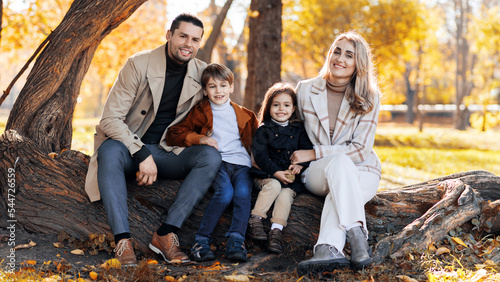 Happy family in an autumn park. Mother, father, son and daughter are sitting on a tree trunk and looking into the camera, yellowed trees around