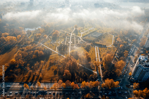 Aerial drone view of Dendrarium Park in Chisinau at sunrise, Moldova. Park full of yellowed trees and grass, fog in the air, residential district around