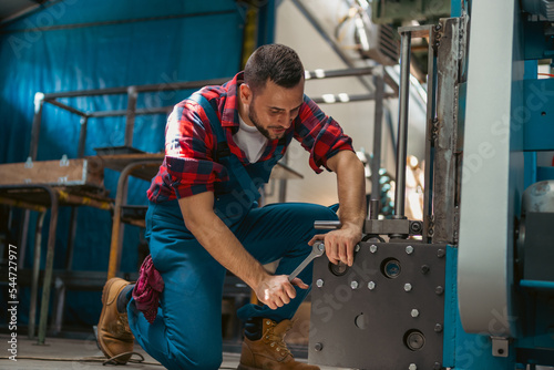 Young male mechanic kneeling on his leg with a smile on his face, while screwing together parts of the metal elements