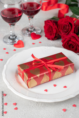 Gift in wrapping paper on white plate, red roses and two glasses wine on light table, concept for Valentines Day
