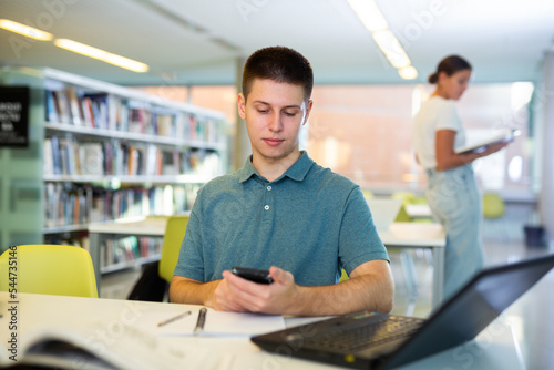 Student young man sitting at a desk in the university library is texting with friends and photographs on a mobile phone