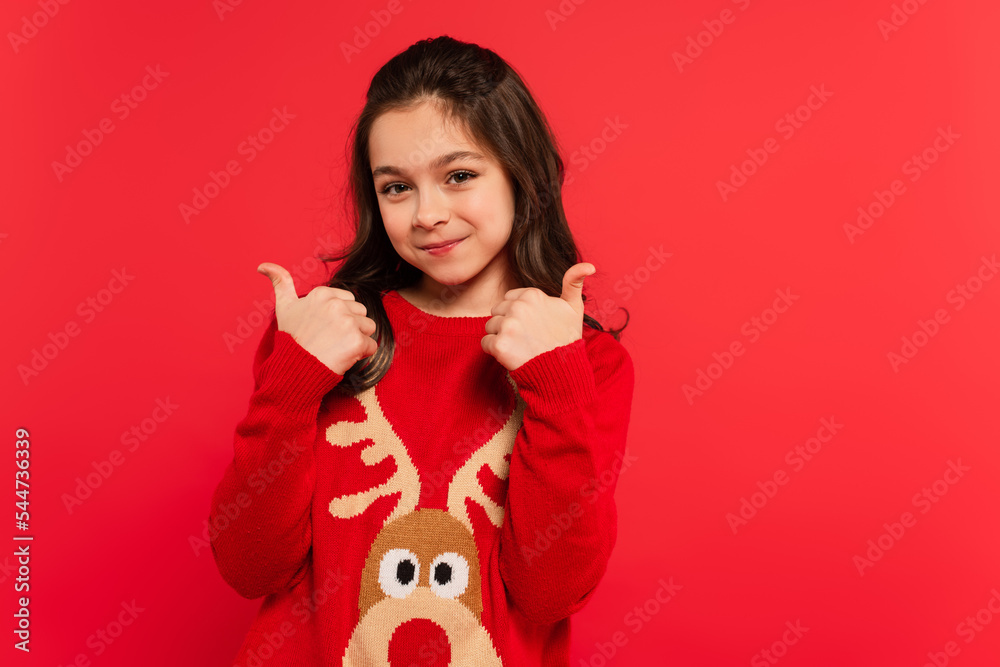 positive girl in winter sweater showing thumbs up and looking at camera isolated on red positive girl in winter sweater showing thumbs up and looking at camera isolated on red