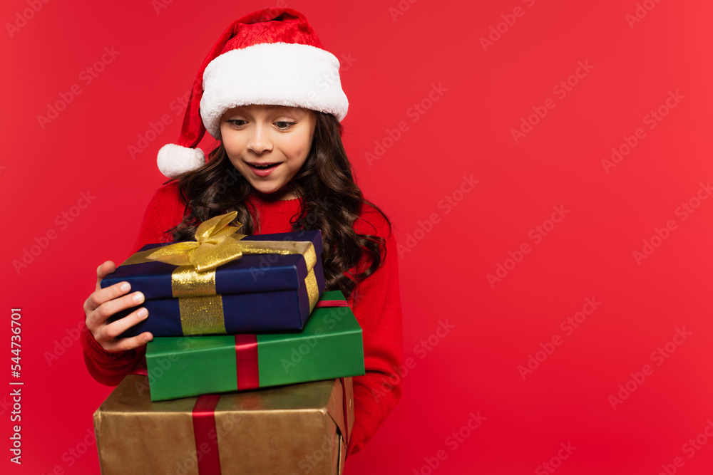 happy girl in santa hat and sweater holding bunch of Christmas presents isolated on red