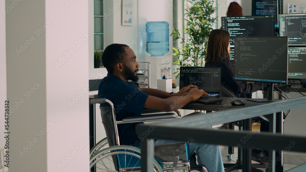 African american IT engineer with disability typing code in it development agency office, using terminal window to write html script database. Wheelchair user working with security programming. African american IT engineer with disability typing code in it development agency office, using terminal window to write html script database. Wheelchair user working with security programming.
