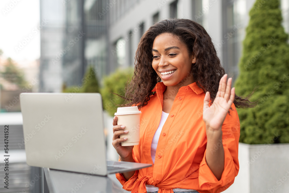 Cheerful millennial black woman with cup of drink takeaway, waving hand at computer webcam, has video call