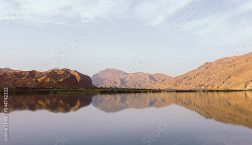 A picturesque panorama of a mountain lake with a reflection at sunrise. A beautiful landscape of a mountain range with a pink pastel sky with hills in the background and reflection in the water.