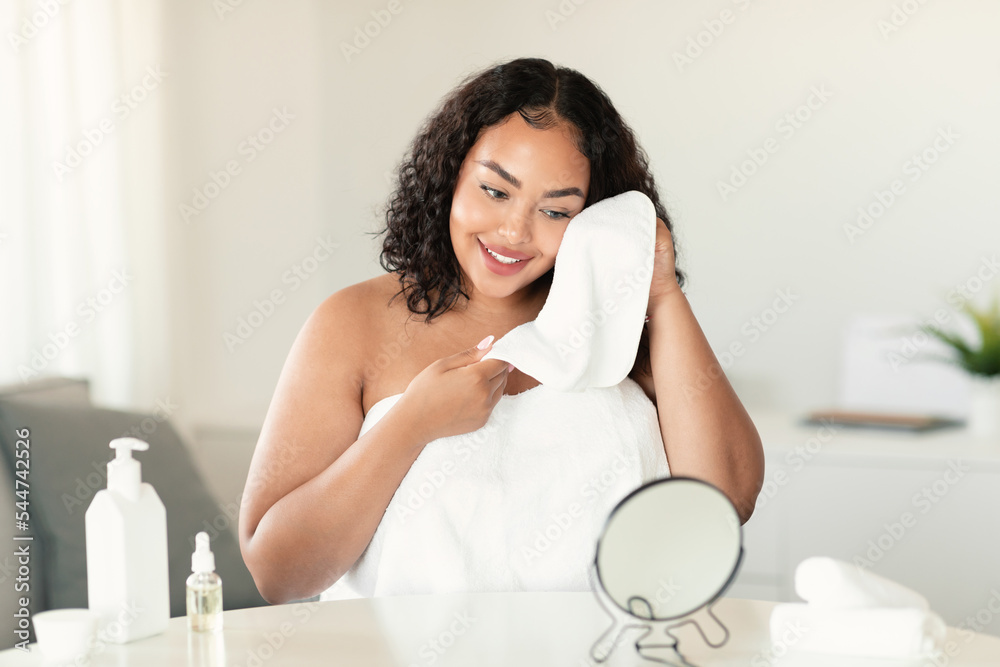 Pretty black chubby woman drying face with soft towel looking at mirror, caring for skin in bedroom interior Pretty black chubby woman drying face with soft towel looking at mirror, caring for skin in bedroom interior