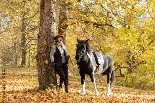 Black and white Icelandic horse and female rider in autumn scenery under maple tree with maple leaf on the ground. Rider wearing black helmet.
