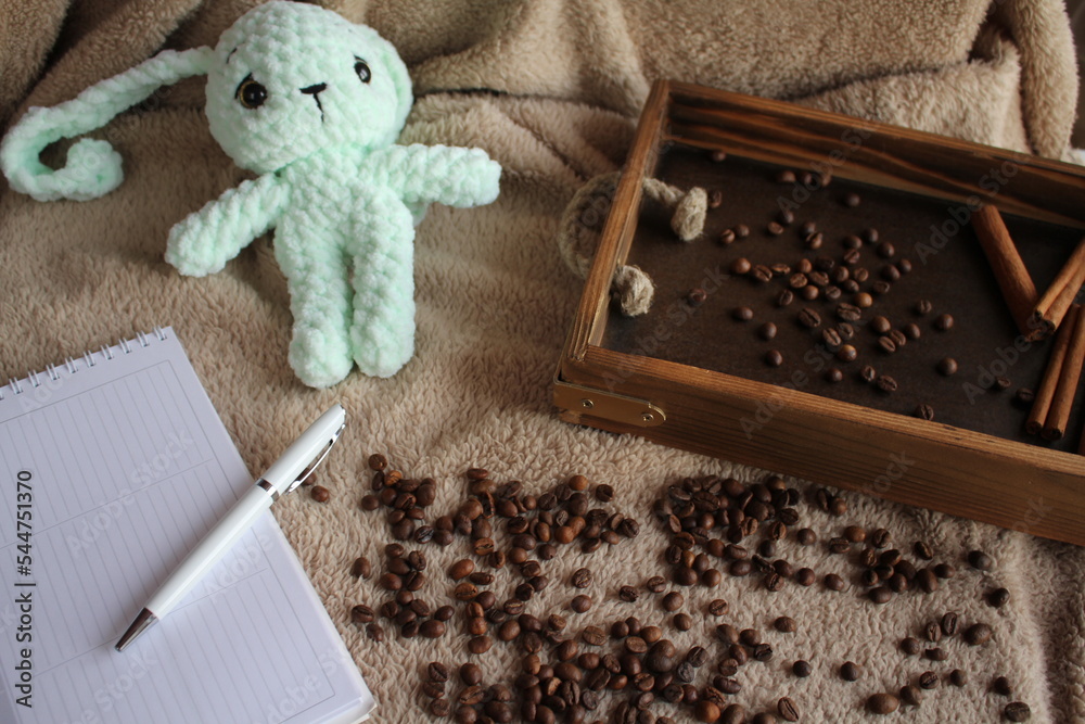 Fototapeta premium A soft toy lies on a table on a plush blanket. There is a wish card next to it. Coffee beans and a wooden tray complete the decor. Handmade. Hobby. Knitting. Gifts for the holidays.