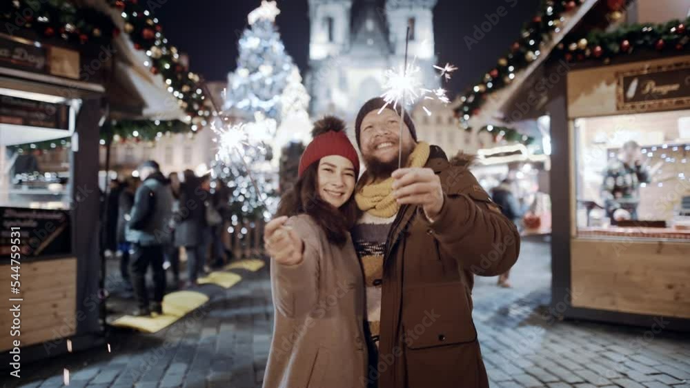 Celebrating New Year holidays vacation. Happy lovely couple with bright sparklers smile at decorated town square. Xmas Christmas winter night atmosphere. Excited people in love kiss having date Celebrating New Year holidays vacation. Happy lovely couple with bright sparklers smile at decorated town square. Xmas Christmas winter night atmosphere. Excited people in love kiss having date