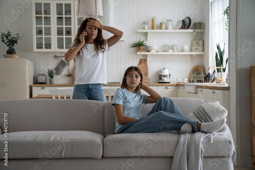 Discouraged woman clutching head talking on phone with daughter teacher and listening to complaints about bad behavior. Frightened teenage girl sits on sofa looking back at mother standing behind