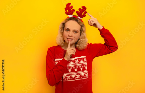 Happy young blonde woman showing silence gesture  pointing to red Christmas reindeer antlers and looking at camera on a yellow background.  Smiling girl wearing knitted red sweater with pattern.