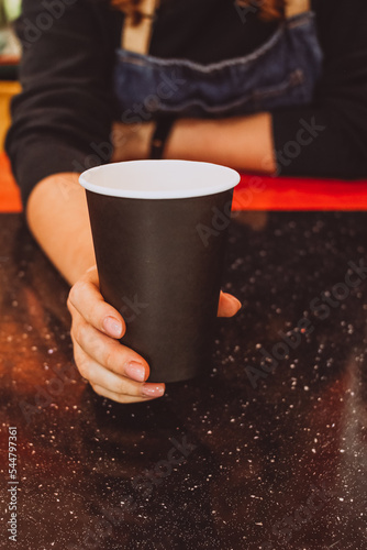 Black cardboard disposable cup in a female hand on a black table, a barista in a coffee shop holds out a glass