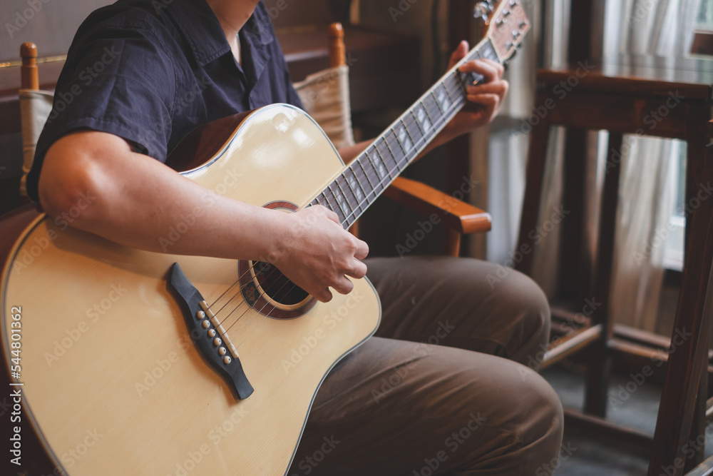 Fototapeta premium Musician is sitting and playing an acoustic guitar for entertainment or practicing for perfomance. Concept of indoor activity, lifestyle and leisure. A guitarist is focusing and enjoy playing guitar.