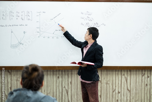 An Asian male teacher in a classroom near the whiteboard is writing and teaching a lesson with a special lecture in a class column. Math teacher explains arithmetic sums to university students photo