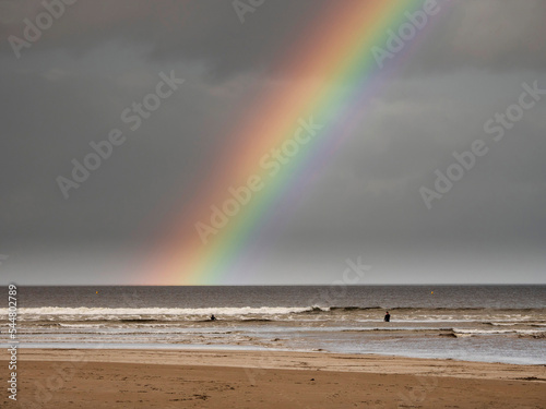 Colorful rainbow in the sky over dark ocean water. Surfers in the waves.