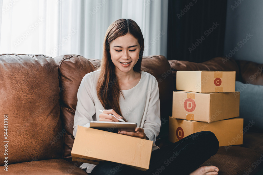 Asian female with small stock business owner holding digital tablet and retail package parcel boxes checking commercial shipping delivery order on smartphone using mobile app technology.