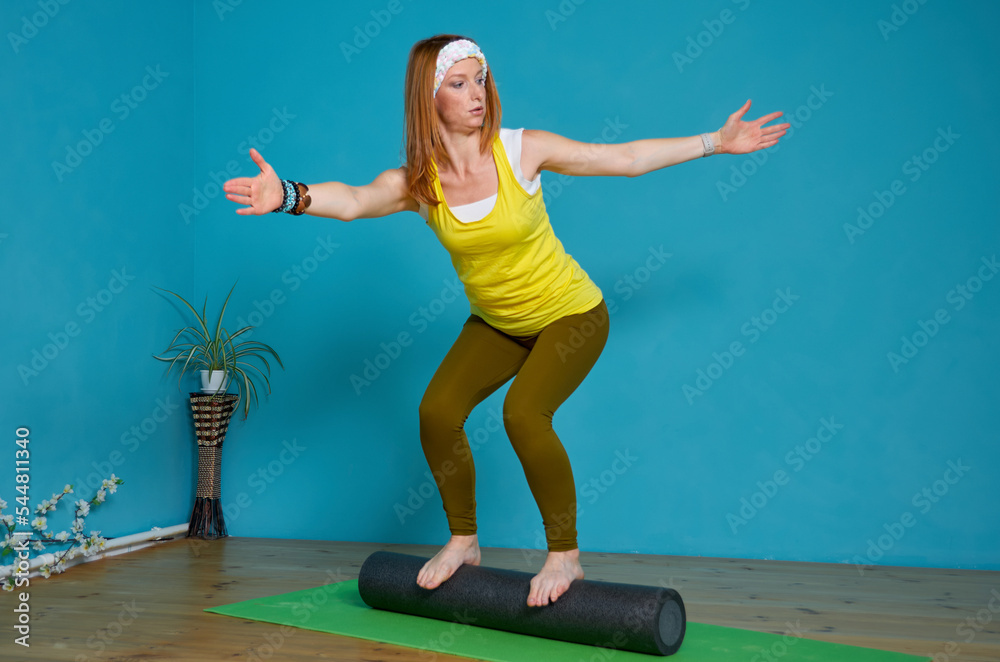 A sporty woman in bright sportswear balances on a roll against a blue wall