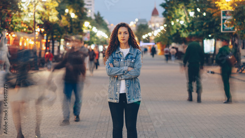 Portrait of stylish young lady tired of usual haste standing in the street among whizzing people and looking at camera. Time, youth and modern society concept.