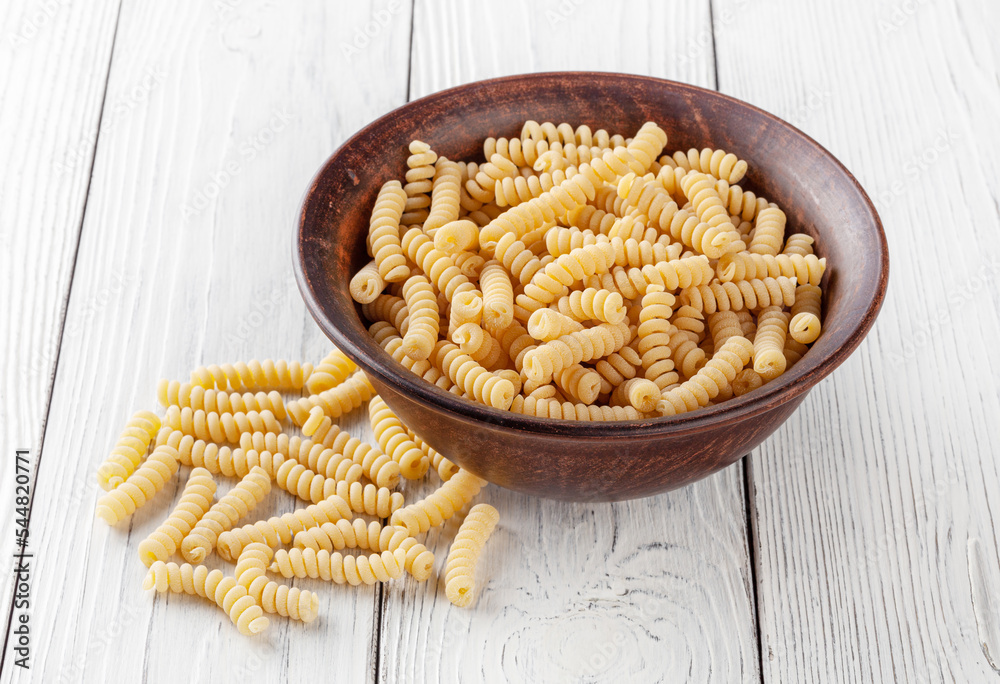 Uncooked fusilli pasta in ceramic bowl on white wooden background