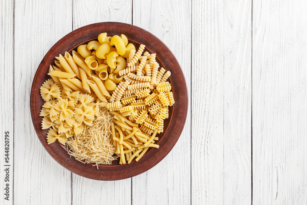 Different types and shapes of Italian pasta in ceramic plate on white wooden background