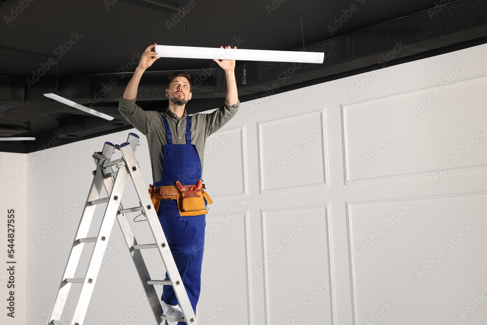 Electrician in uniform installing ceiling lamp indoors. Space for text