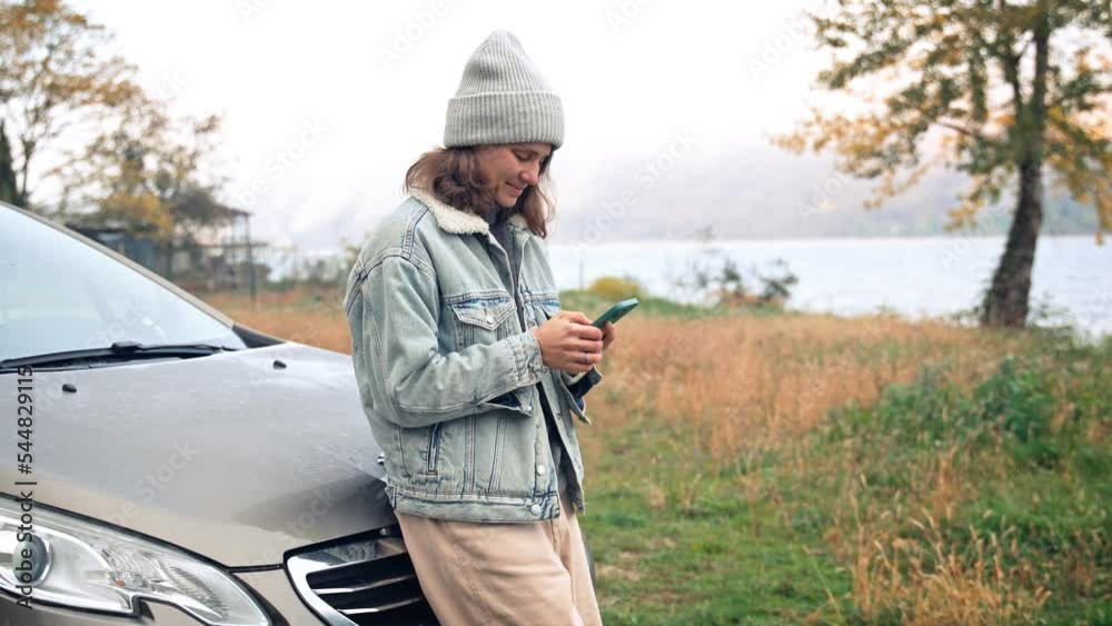 A young caucasian woman is using her smartphone and enjoying a beautiful view of the mountains while leaning on the car hood. A young caucasian woman is using her smartphone and enjoying a beautiful view of the mountains while leaning on the car hood.