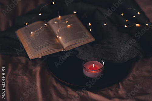 Open peper book with knitted cloth textile and scented burning candle in glass jar on wooden tray in bed on pink duvet close up at night. Winter holiday season.