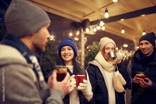 Happy smiling friends with cups of mulled wine having fun, spending time together at winter fair at evening time. Winter holidays, Christmas concept