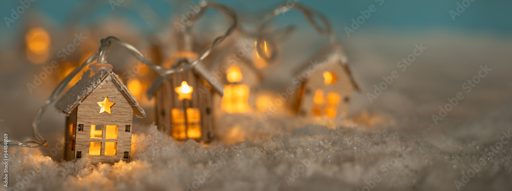 Obraz premium Abstract Christmas Winter Panorama with Wooden Houses Christmas String Lights in Cold Snow Landscape and Glowing Golden Lights in Background. Panorama, Banner. Christmas or Energy themes.