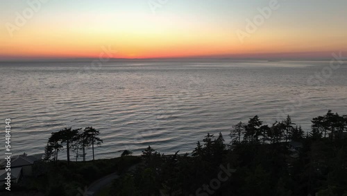 Aerial view pulling away from the sunset to reveal a forest and homes overlooking the water. photo