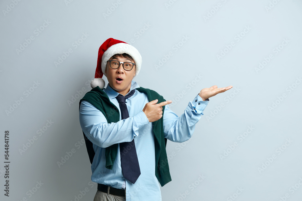 Shocked Man Pointing Aside. Astonished Asian Guy in Santa Claus Hat Pointing at Copy Space and Looking With Amazement. Indoor Studio Shot Isolated on Grey Background 