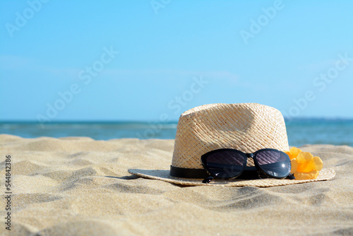 Hat with beautiful sunglasses and flowers on sand near sea. Space for text