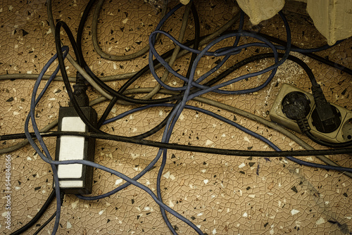 Electrical plugs with cords connected to power strip. Cable power cords in a tangled mess on floor of workplace, used dirty electric outlet