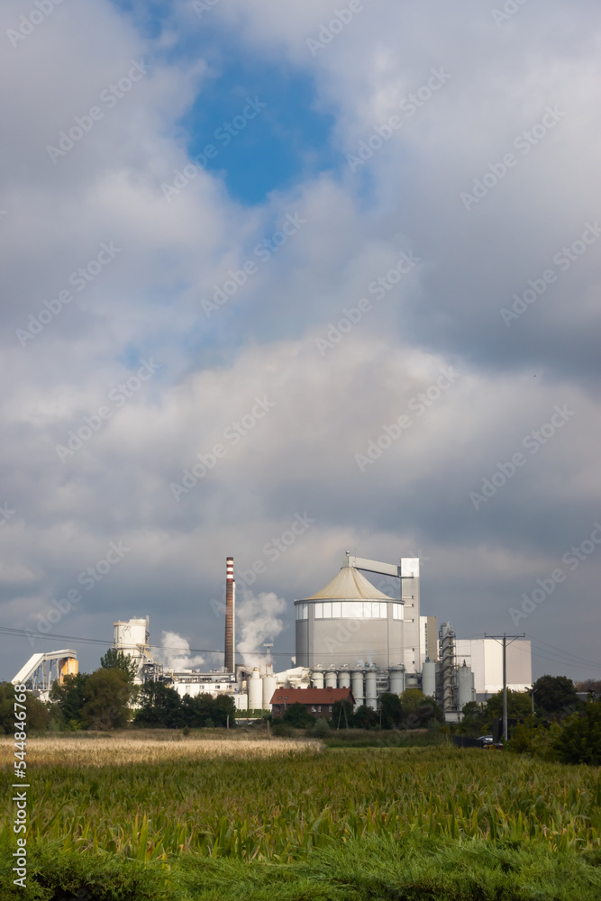 A large industrial plant in the distance. Sugar factory against the slightly cloudy sky. Photo taken in natural lighting conditions.