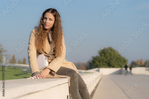 Close-up of thoughtful mid adult woman with curly hair looking in camera at park. Dreamy woman in beige coat autumnal park.