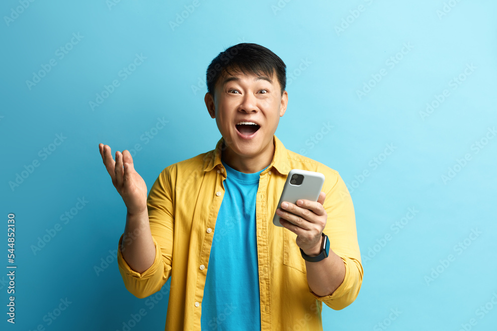 Winner Man Rejoicing Smartphone. Extremely Happy Asian Guy Raised Arm Gesturing Yes I Did It, I'm Winner, Feeling Energetic and Lively. Isolated on Blue Background Winner Man Rejoicing Smartphone. Extremely Happy Asian Guy Raised Arm Gesturing Yes I Did It, I'm Winner, Feeling Energetic and Lively. Isolated on Blue Background